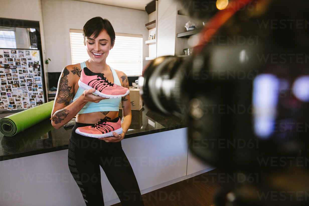 A female influencer is filming herself while holding up a pair of orange shoes and smiling at the camera. 